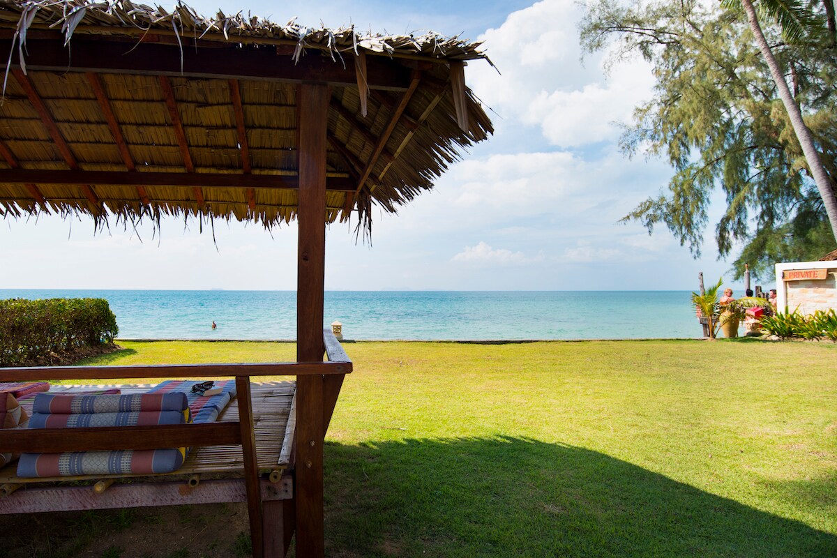An outdoor seating area features a thatched-roof pavilion overlooking the calm sea. Lush green grass extends to the shoreline, with a serene view of the water and distant horizon under a partly cloudy sky.