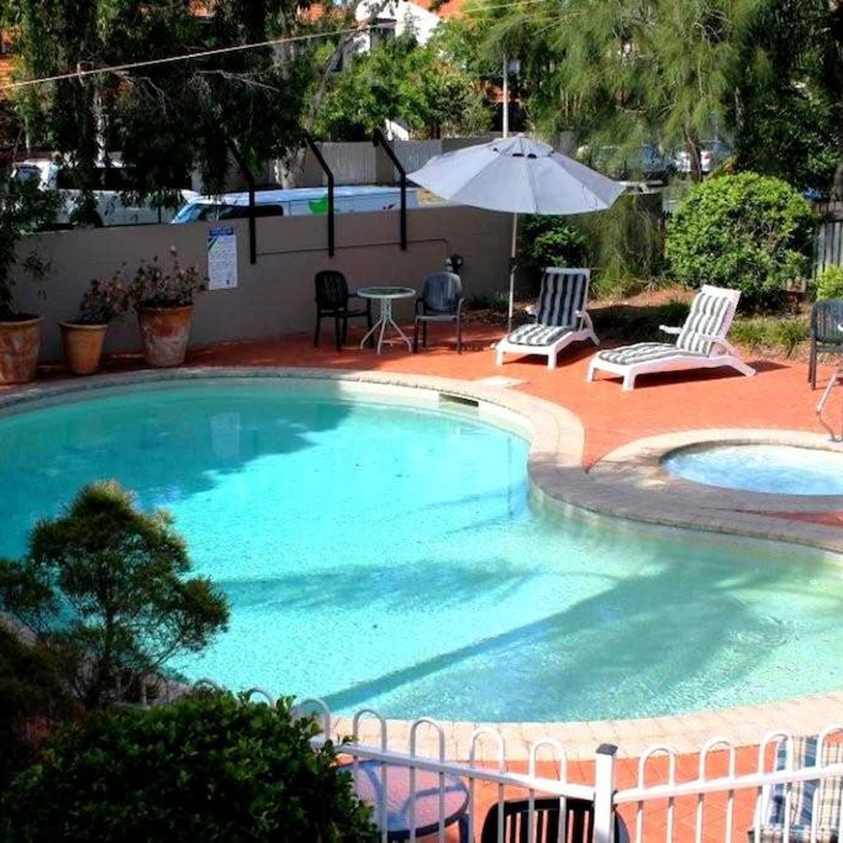 A sparkling pool is surrounded by lush greenery and arranged with comfortable lounge chairs and a shaded table. A small hot tub is visible adjacent to the pool, providing a relaxing element in this inviting outdoor space.