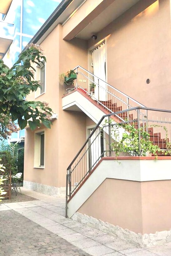 A two-story building is shown with a light-colored exterior. An exterior staircase leads to a small landing, adorned with potted plants. The pathway is paved, bordered by greenery and a seating area visible to the side.