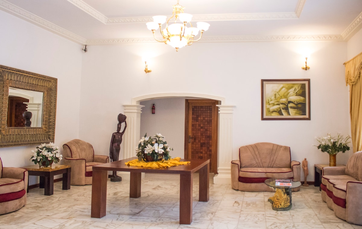 A spacious living area features elegant furnishings, including two plush armchairs and a central wooden table. Decorative plants are arranged on a side table, and a wall art piece complements the decor. Mirrored designs and a statue add a cultural touch to the ambiance.