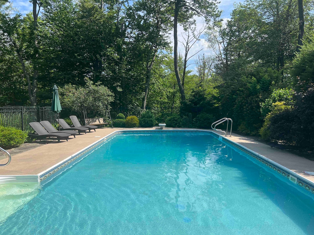 A serene pool area is surrounded by lush greenery, creating a private retreat. Lounge chairs are positioned nearby, providing a place to relax. A tall umbrella stands ready to offer shade during sunny days, enhancing the tranquil setting.