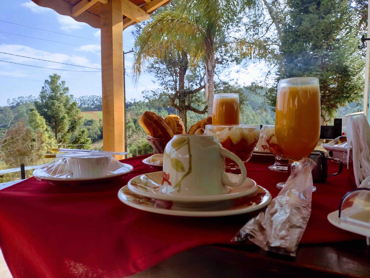 A breakfast table is set with a variety of foods, including pastries, fruit, and drinks. A scenic view of green hills is visible in the background, framed by trees. The setting features warm sunlight illuminating the table arrangement.