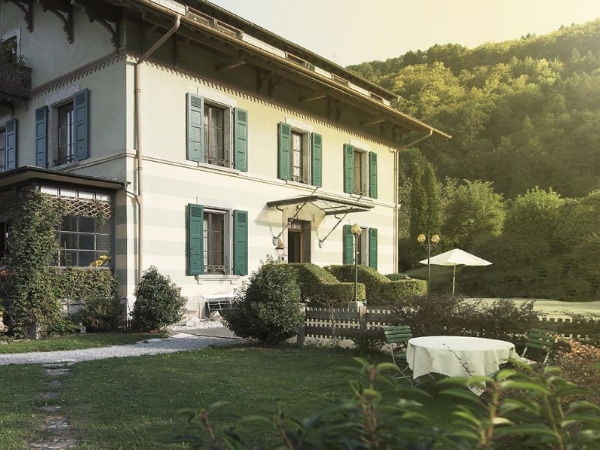 The exterior of the building is characterized by vibrant green shutters and a welcoming entrance. Lush green landscaping surrounds the property, with a small table set on the grassy area, offering a tranquil outdoor space. A shaded umbrella provides comfort on sunny days.