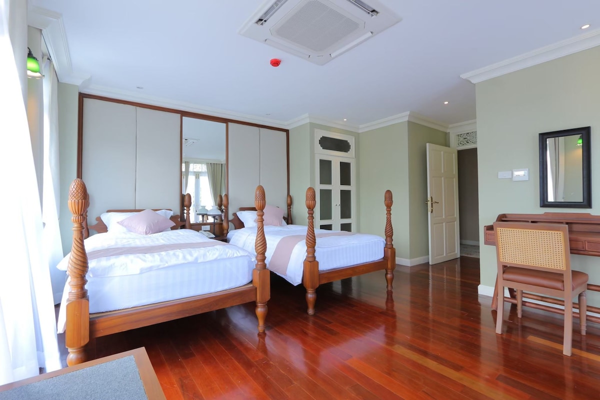 A spacious room features two separate single beds with classic wooden frames. Natural light enters through large windows with sheer curtains. A wooden desk and mirror are positioned neatly against the wall, while polished hardwood floors add warmth to the space.