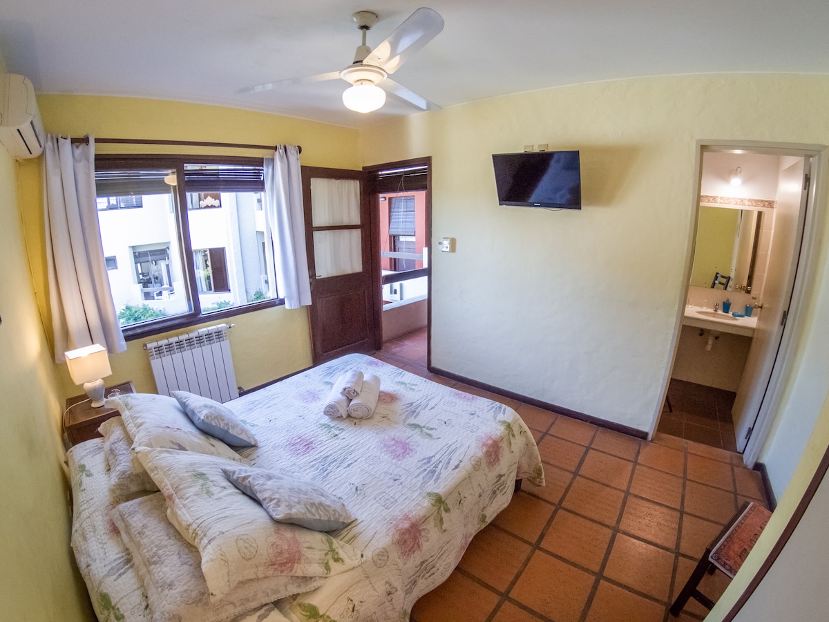 A private room features a spacious bed with light-colored bedding adorned with floral patterns. A ceiling fan and wall-mounted television are visible. Natural light enters through the windows, and an en-suite bathroom is seen through an open door. Tile flooring adds texture to the space.