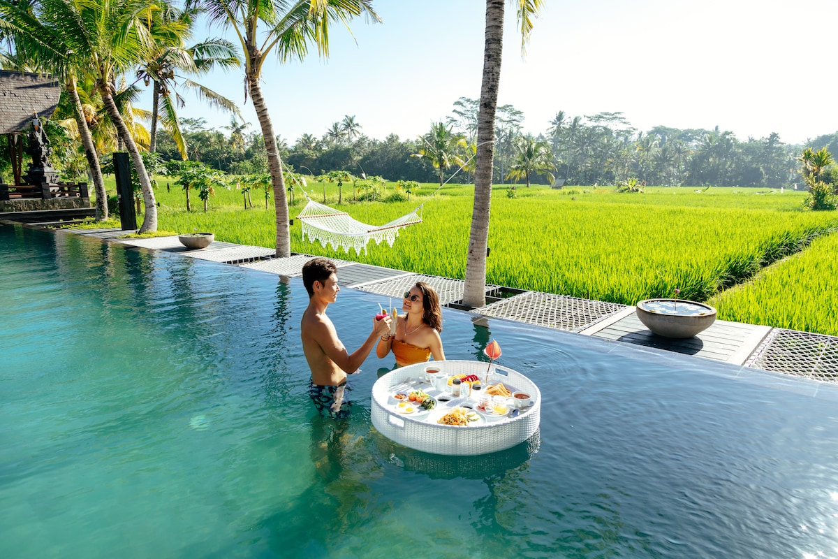 A couple enjoys refreshments while seated in a floating tray within an infinity pool. Lush green rice fields stretch into the distance, framed by palm trees. A hammock is visible on the edge of the property, enhancing the tranquil atmosphere.