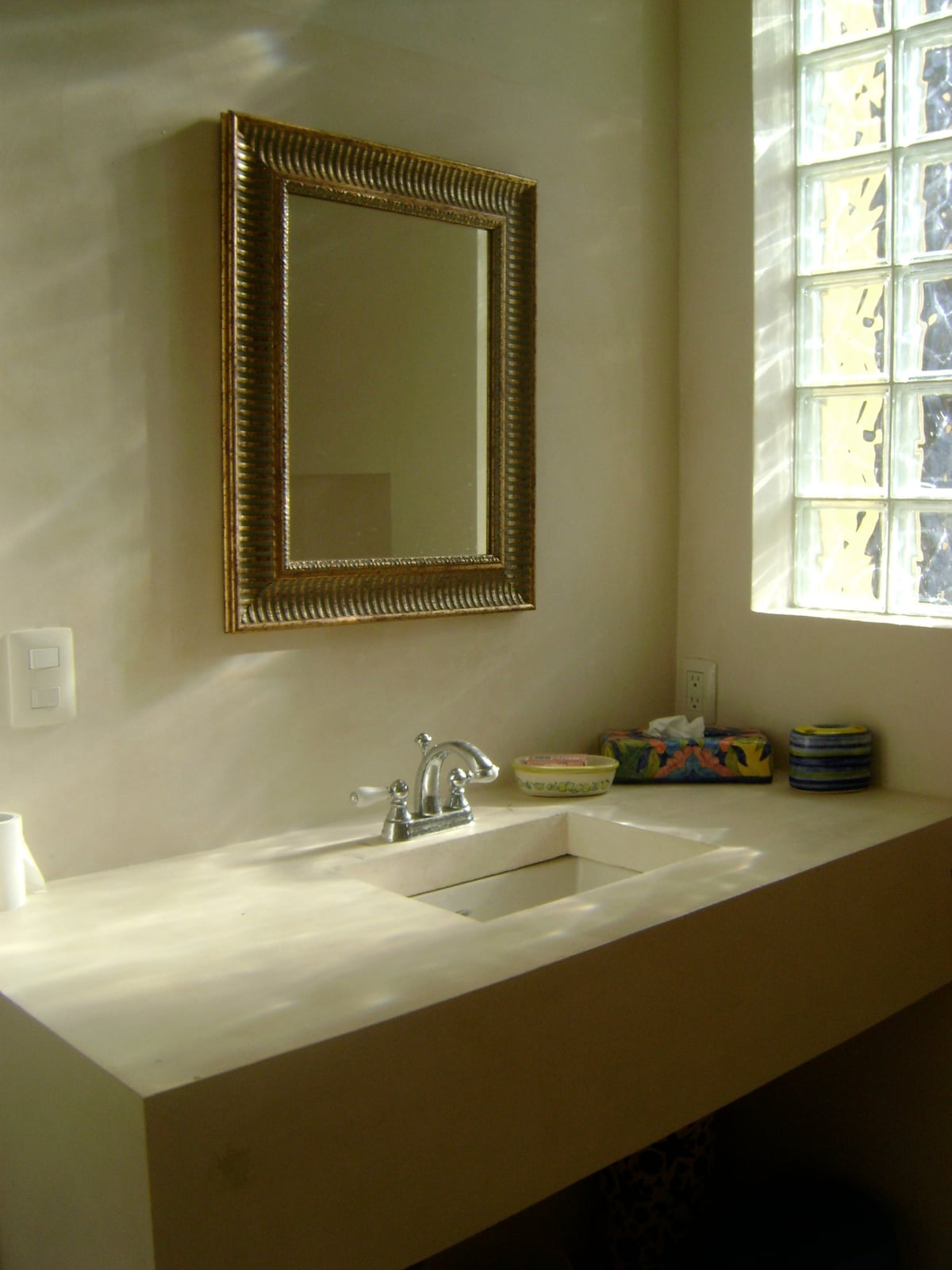 A well-lit bathroom features a sleek countertop with a built-in sink. A decorative mirror hangs above, reflecting light. Natural light enters through a glass block window, accentuating the neutral color scheme. Toiletries and decorative items are neatly arranged nearby.