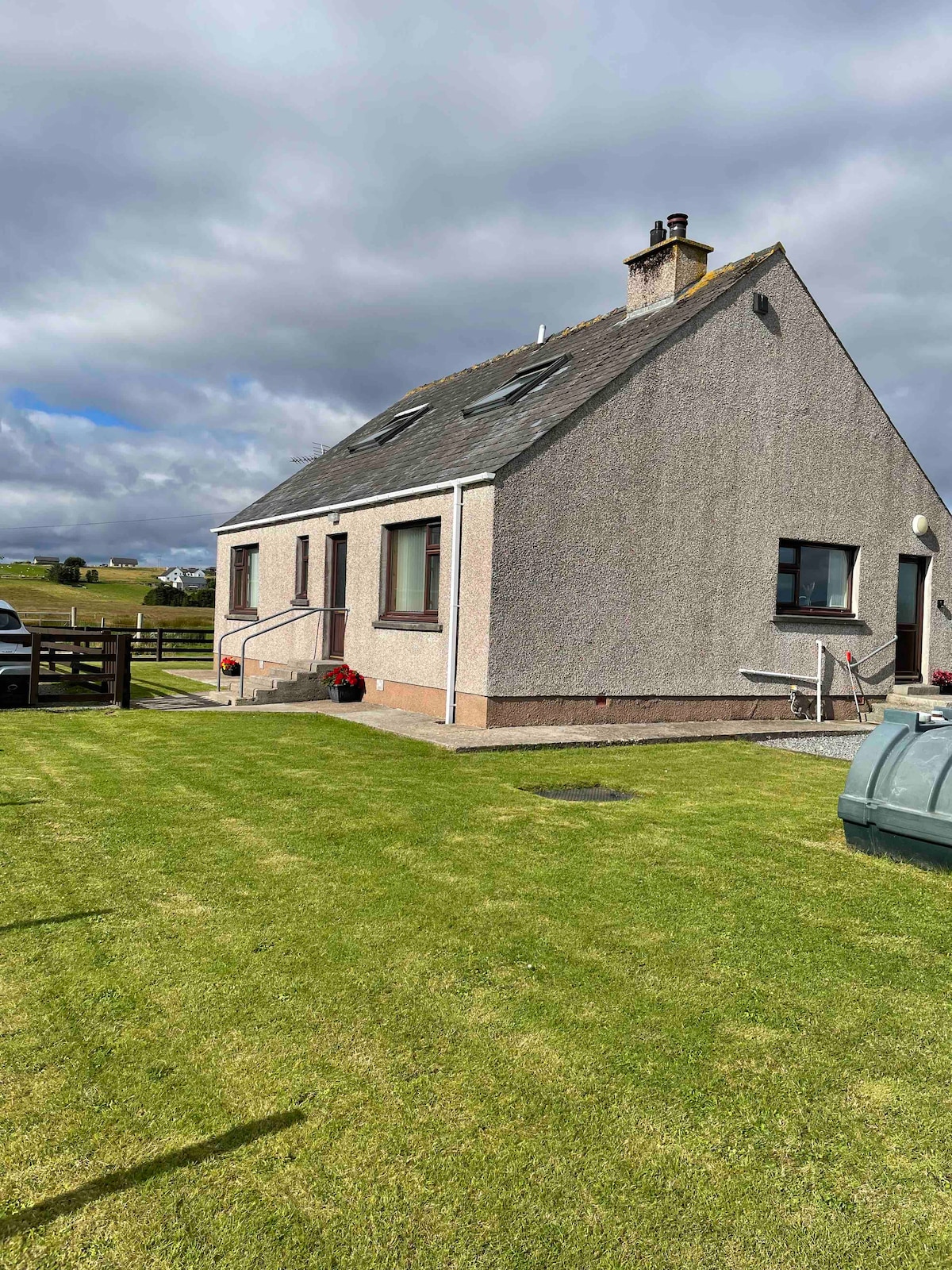 The image captures the exterior of a detached two-story holiday home with a sloped roof, set against a cloudy sky. A well-maintained lawn surrounds the property, while a small patio area and steps lead to the entrance. A glimpse of the surrounding landscape is visible.