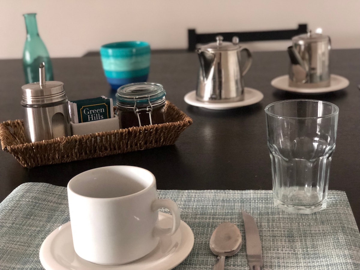 A dining table is set with a woven tray holding a sugar container and a teapot. A clear glass and a white cup are positioned on a light blue placemat. Additional tableware includes a silver container and assorted decorative bowls in calming hues.