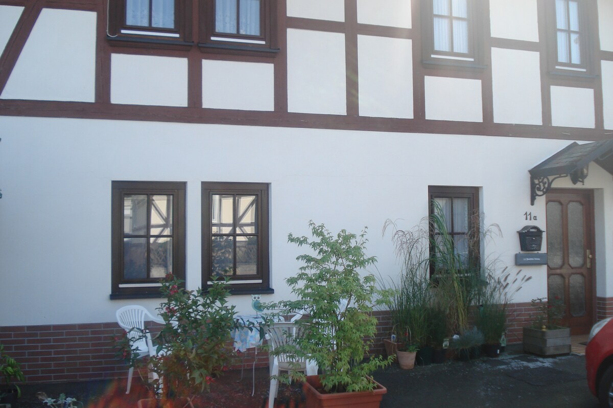The exterior of a half-timbered building is visible, featuring two small windows and a decorative entryway. A seating area with white chairs and a table sits in front, surrounded by potted plants and greenery, adding a touch of nature to the entrance.