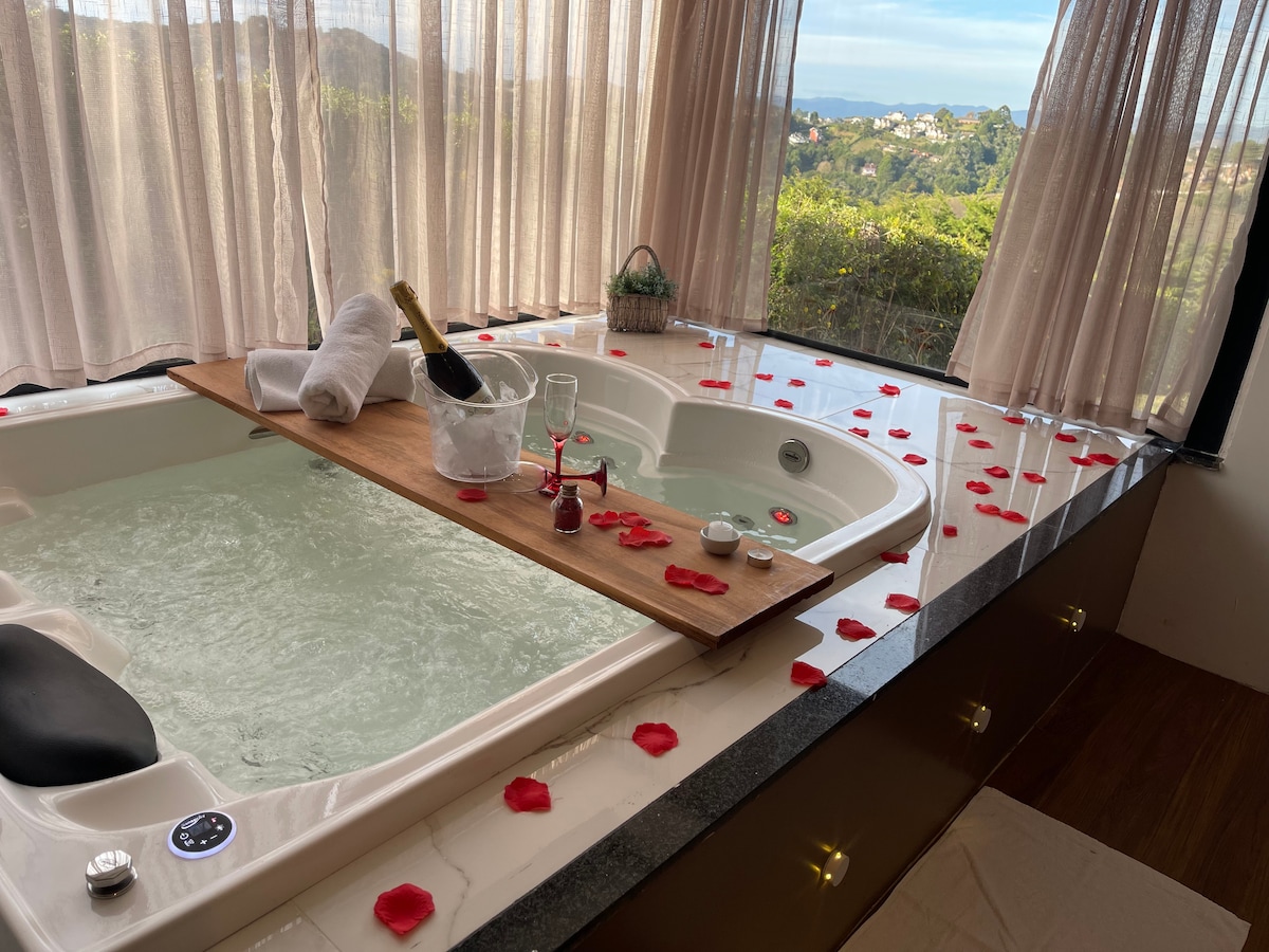 A spacious whirlpool bath is positioned near a large window, offering a scenic view of the outdoors. The surrounding countertop is adorned with rose petals and a decorative bottle, creating a serene and romantic atmosphere.