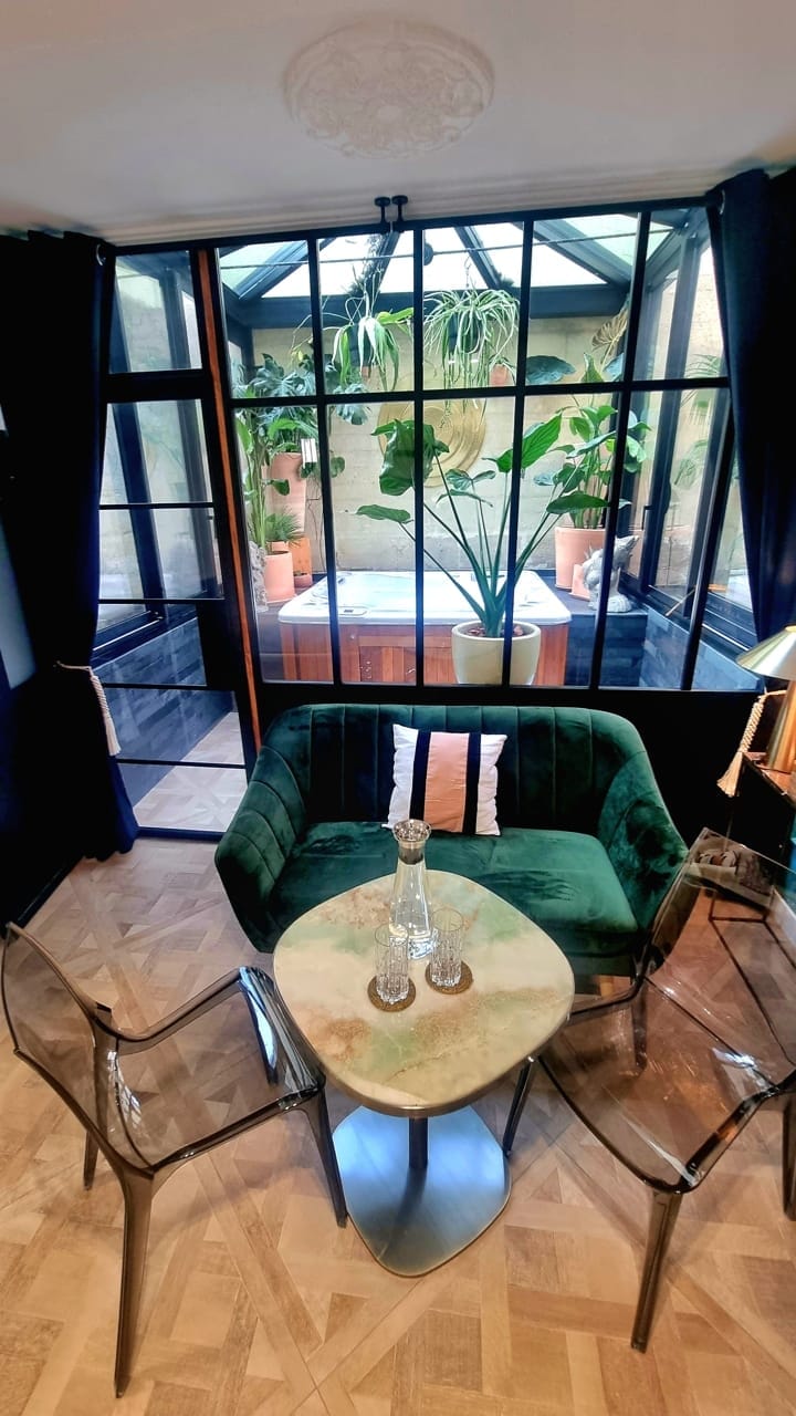 A bright seating area is presented with a green velvet couch and transparent chairs around a marble table. Large windows frame a view of an outdoor space filled with greenery. Light pours in, enhancing the fresh atmosphere of the room.