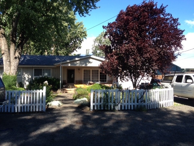The exterior of a single-story home is seen, framed by a white picket fence. A lush leafy tree provides shade in the front yard, while a gravel driveway leads to the entrance, where porch seating is visible. A clear blue sky complements the scene.