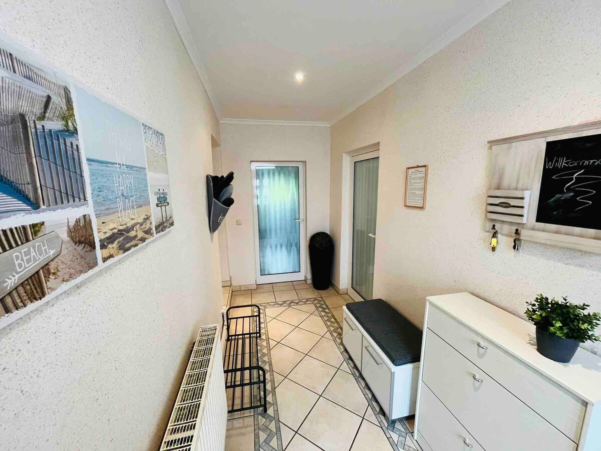 A bright and inviting hallway features light-colored walls and tiled flooring. Beach-themed artwork is displayed on one side. A storage bench is positioned against the wall, alongside a white cabinet. A door leads to an outdoor space, with additional doors visible in the background.