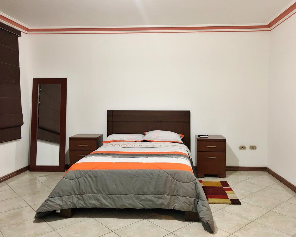 A spacious sleeping area features a large bed with a grey and orange quilt. Two wooden nightstands are positioned on either side of the bed, and a mirror is mounted against the wall. Natural light enters through a window with brown blinds, illuminating the tiled floor.