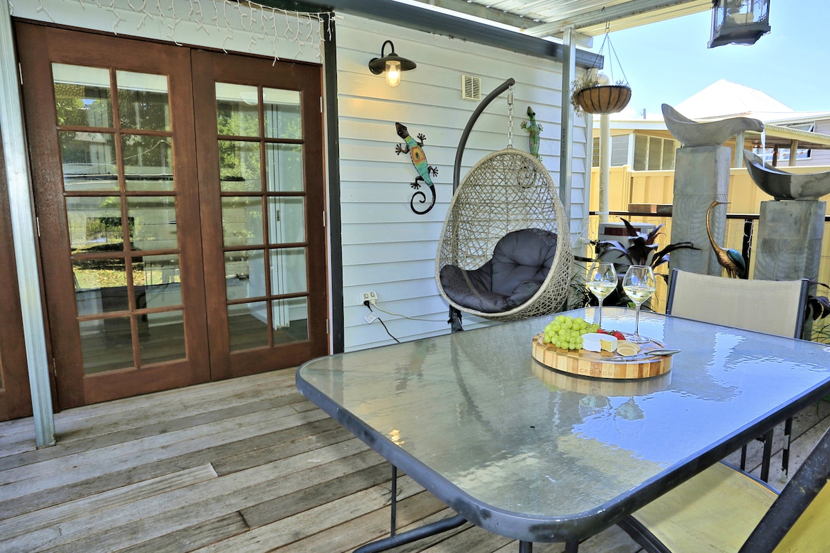 An outdoor dining area features a glass table set with decorative items and glasses. A cozy hanging chair invites relaxation, while double French doors reveal a glimpse of indoor space. Natural wood flooring adds warmth to the area, surrounded by greenery.