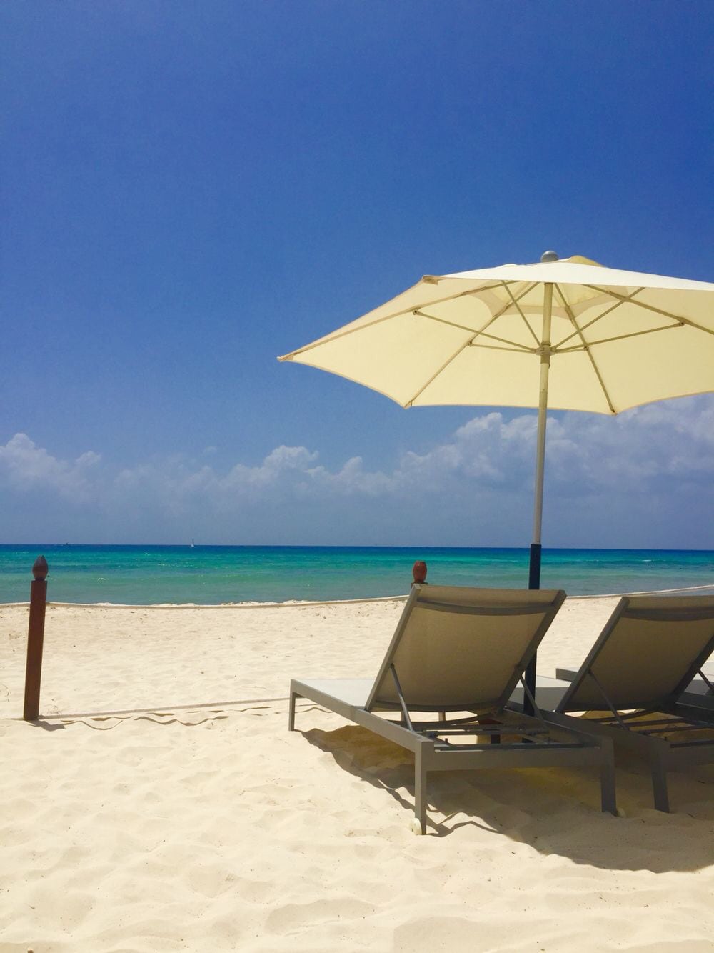 A serene beach setting is depicted, featuring two comfortable lounge chairs shaded by a large umbrella. The white sand extends towards a tranquil sea, showcasing varying shades of blue and a clear sky with soft clouds in the background.