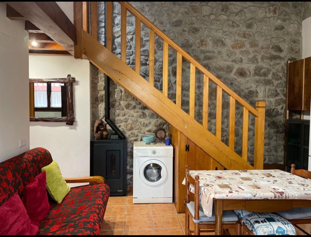 A living area features a wooden staircase leading to an upper level, flanked by a rustic stone wall. A red-patterned sofa sits beside a compact dining table with wooden chairs, while a washing machine is positioned near a heating stove.