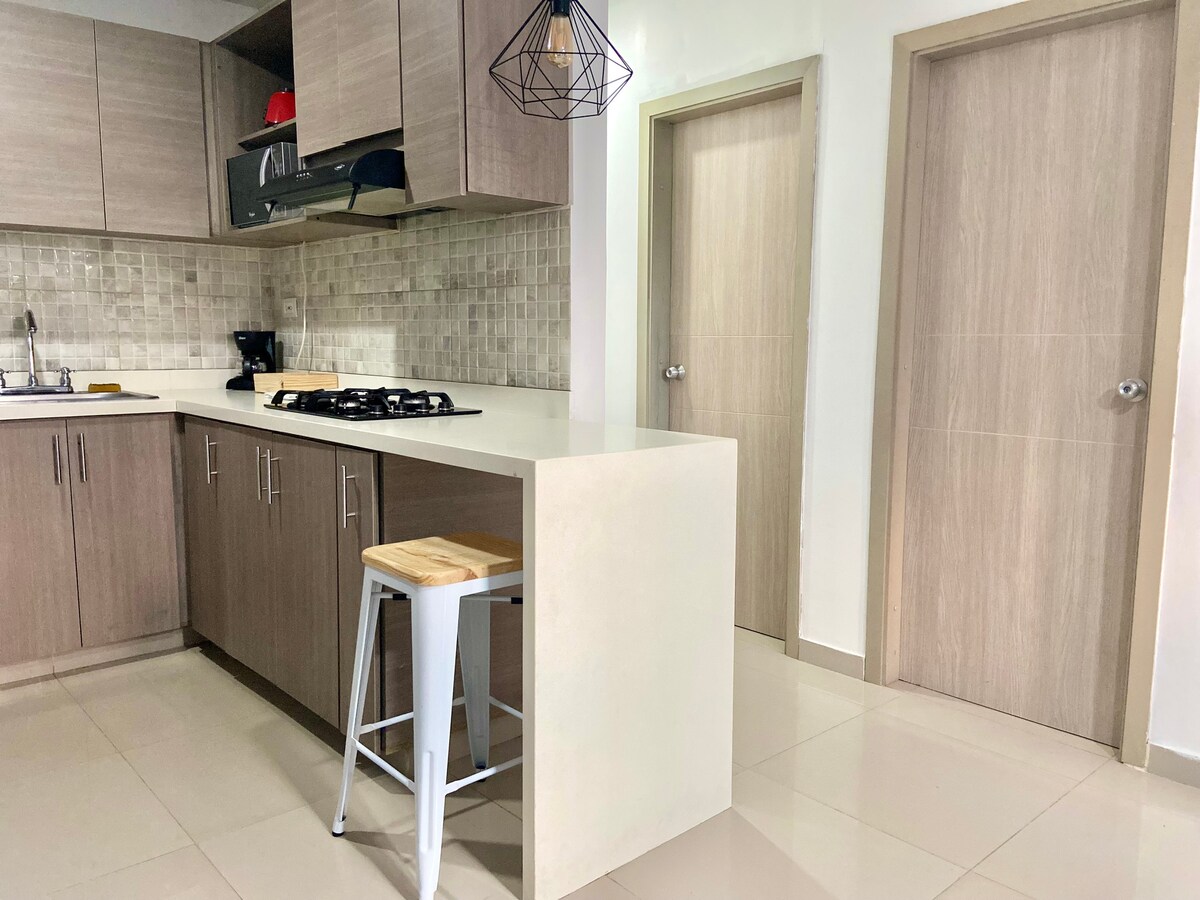 A modern kitchen is featured with light wooden cabinetry and a neutral backsplash. An island with a built-in stove is complemented by a metal bar stool. Two closed doors are visible, leading to additional rooms, and the space is well-lit with artificial lighting.