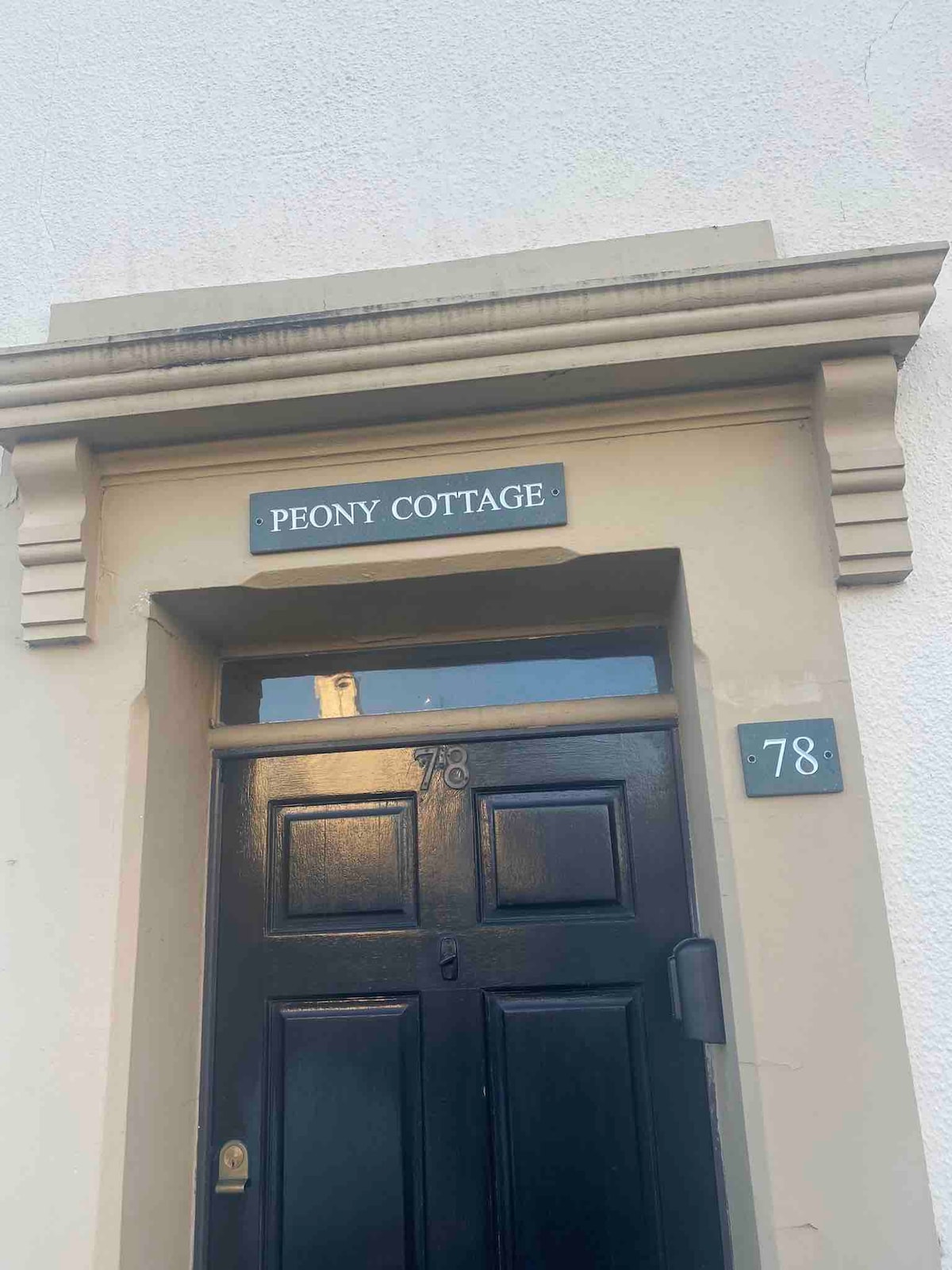 The entrance of Peony Cottage is showcased with a dark wooden door featuring a classic design. A plaque with 'PEONY COTTAGE' is displayed above the door, while the number '78' is positioned beside it. Light reflects gently off the door surface.