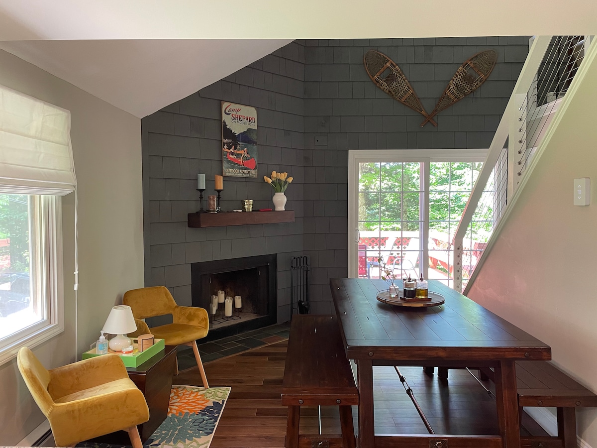A cozy dining area is featured with a large wooden table and yellow accent chairs. A fireplace is positioned against a gray wall, accompanied by decorative items and candles. Natural light filters in through large windows, revealing a view of the surrounding greenery.
