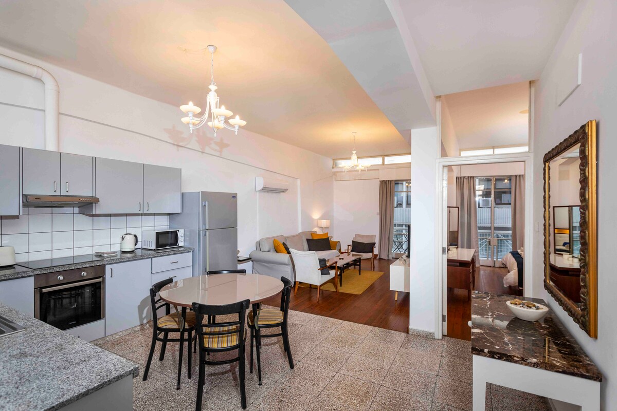 The open-plan living area showcases a dining table with four chairs, situated beside a fully equipped kitchen. The comfortable seating area includes a sofa and armchair, illuminated by natural light from the balcony doors. Decorative elements like a mirror and artwork add to the ambiance.