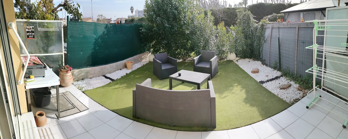 An outdoor garden area is visible, featuring a small seating arrangement with two armchairs and a table on a green lawn surface. A planter is placed on the ground, and the area is surrounded by greenery, enhancing the space's tranquility. A washing line stands in the corner.