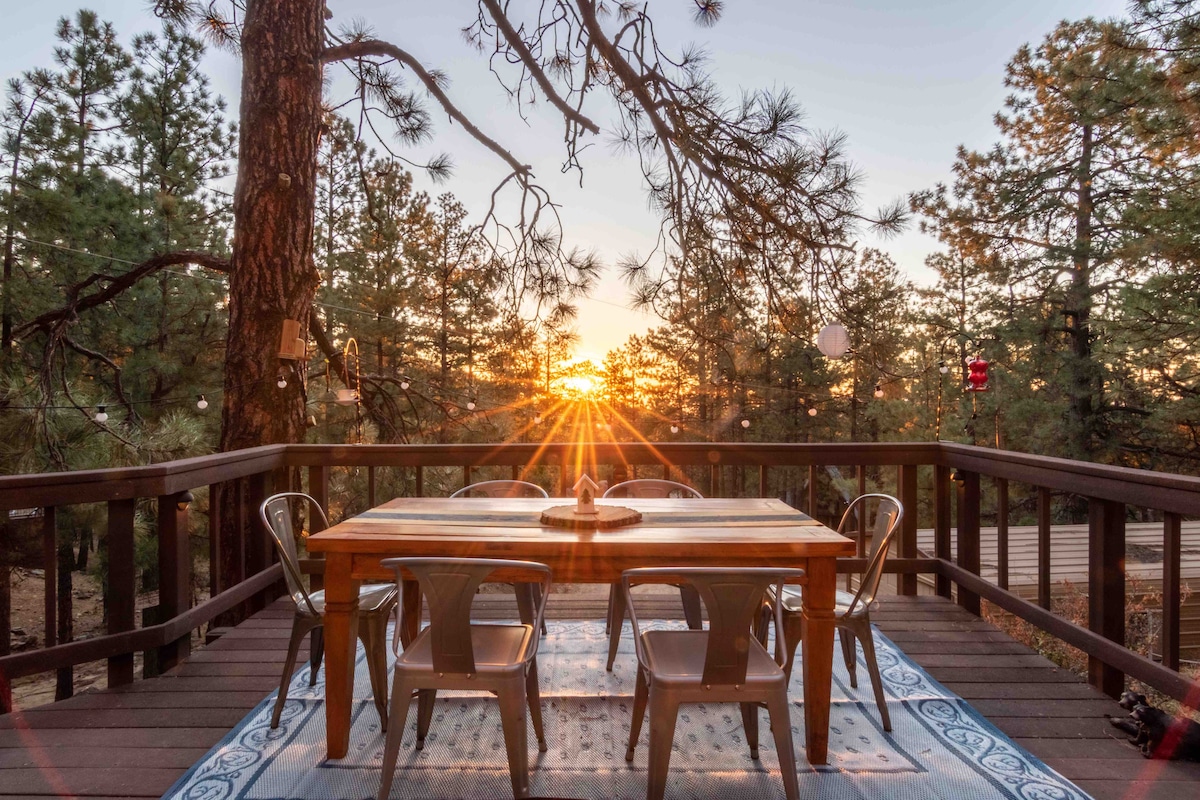 A wooden deck features a rectangular dining table surrounded by six metal chairs. The scene captures a sunset filtering through pine trees, casting a warm glow across the area. A patterned rug lies beneath the table, enhancing the outdoor dining setting.