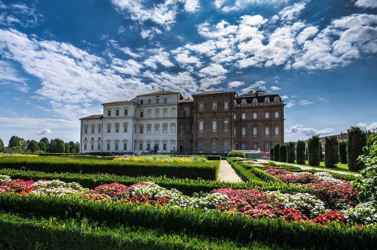 A grand historical building is presented with a blend of light-colored and brick exteriors. Lush landscaped gardens filled with colorful flowers and neatly trimmed hedges surround the structure, set against a bright blue sky with scattered clouds.