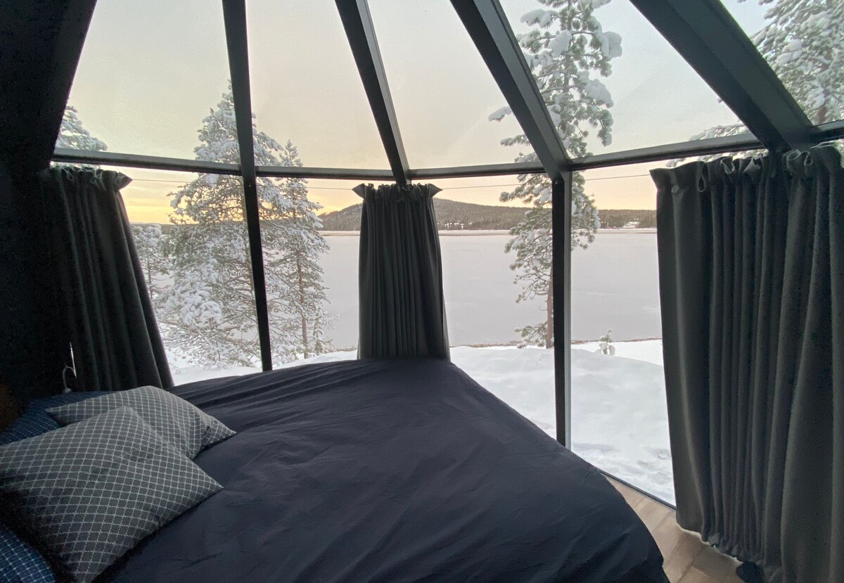 A spacious sleeping area features large glass windows offering views of a snow-covered landscape. Soft curtains frame the windows, complementing the minimalist decor. The bed is neatly made with dark bedding and decorative pillows, providing a comfortable atmosphere close to nature.