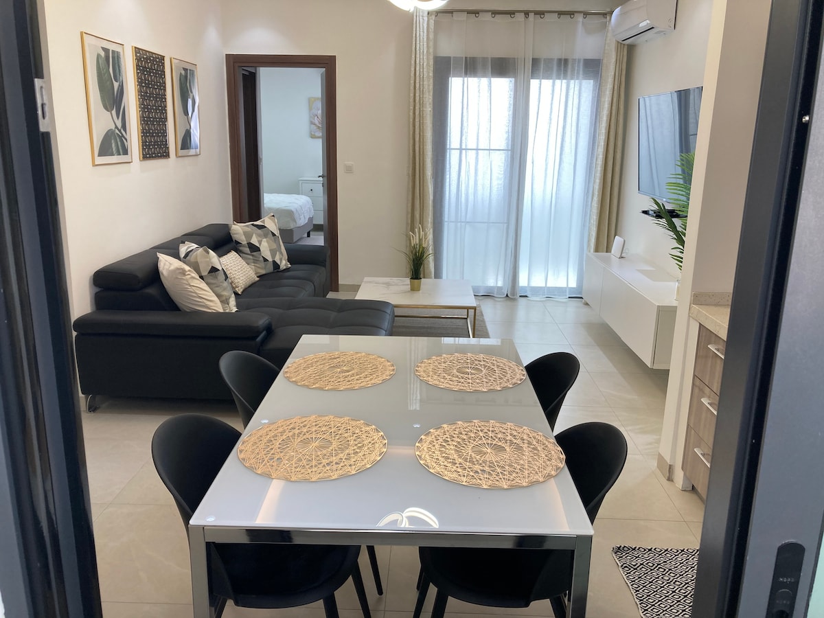 A modern living space features a sleek black sofa paired with decorative cushions. A dining table for six is set with woven placemats. Large windows allow natural light to fill the room, creating a seamless transition to a bright area with additional furnishings.