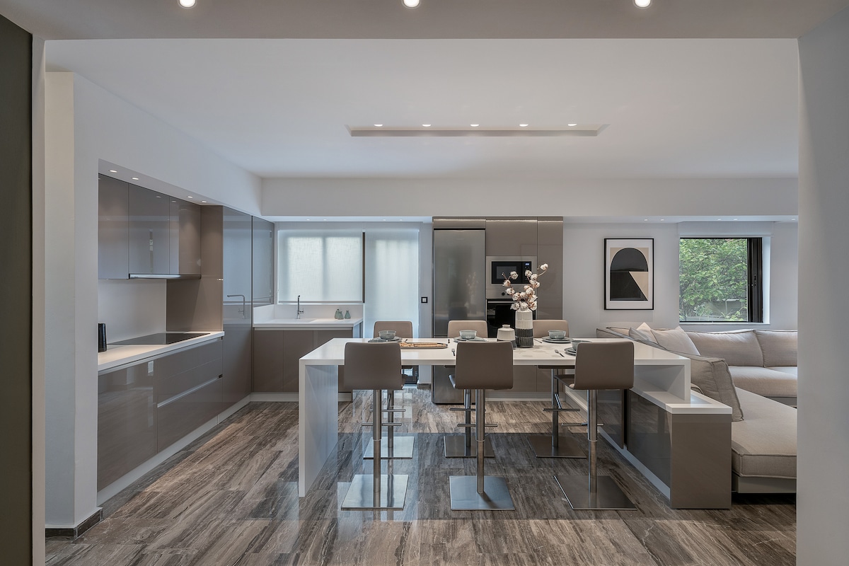 A modern kitchen and dining area are showcased, featuring sleek cabinetry and a spacious island set with four dining chairs. Natural light fills the room through large windows. A comfortable sofa is positioned nearby, providing a relaxed seating area.
