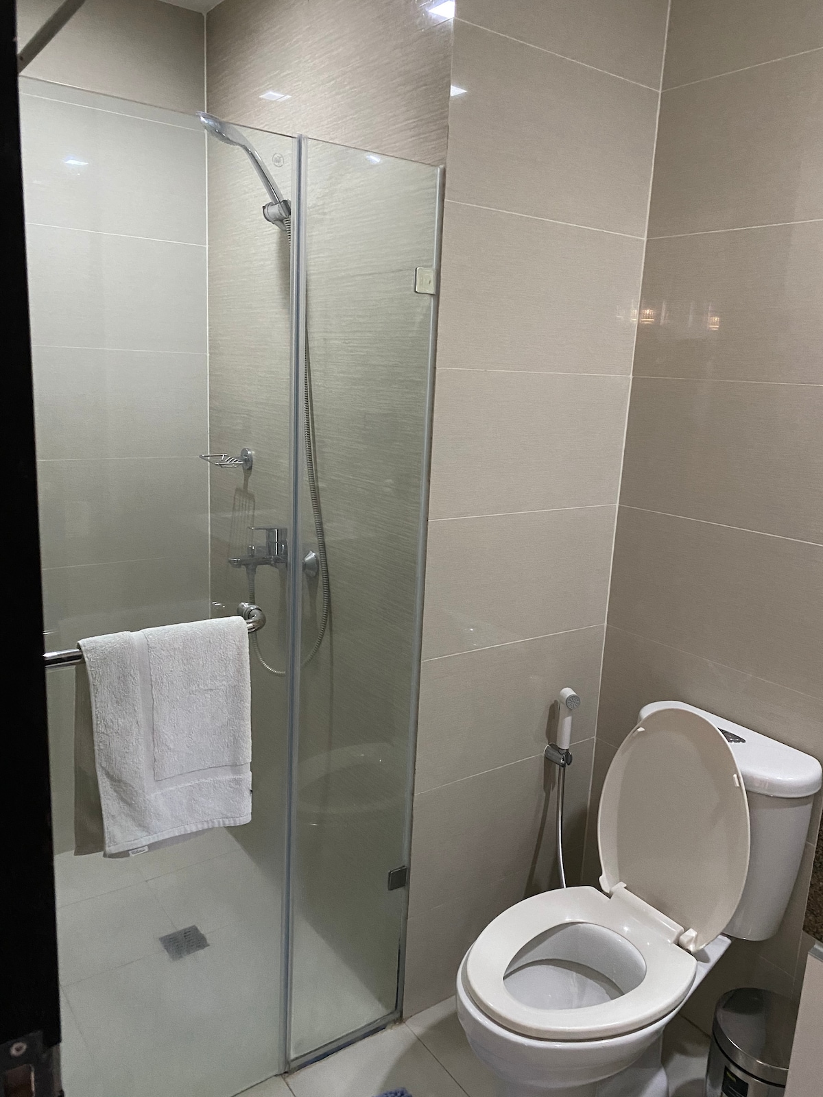A modern bathroom is depicted, featuring a glass-enclosed shower with a sleek showerhead. A white towel hangs on a chrome bar beside the shower. A clean toilet is positioned next to a wastebasket, with neutral tiles providing a cohesive look.