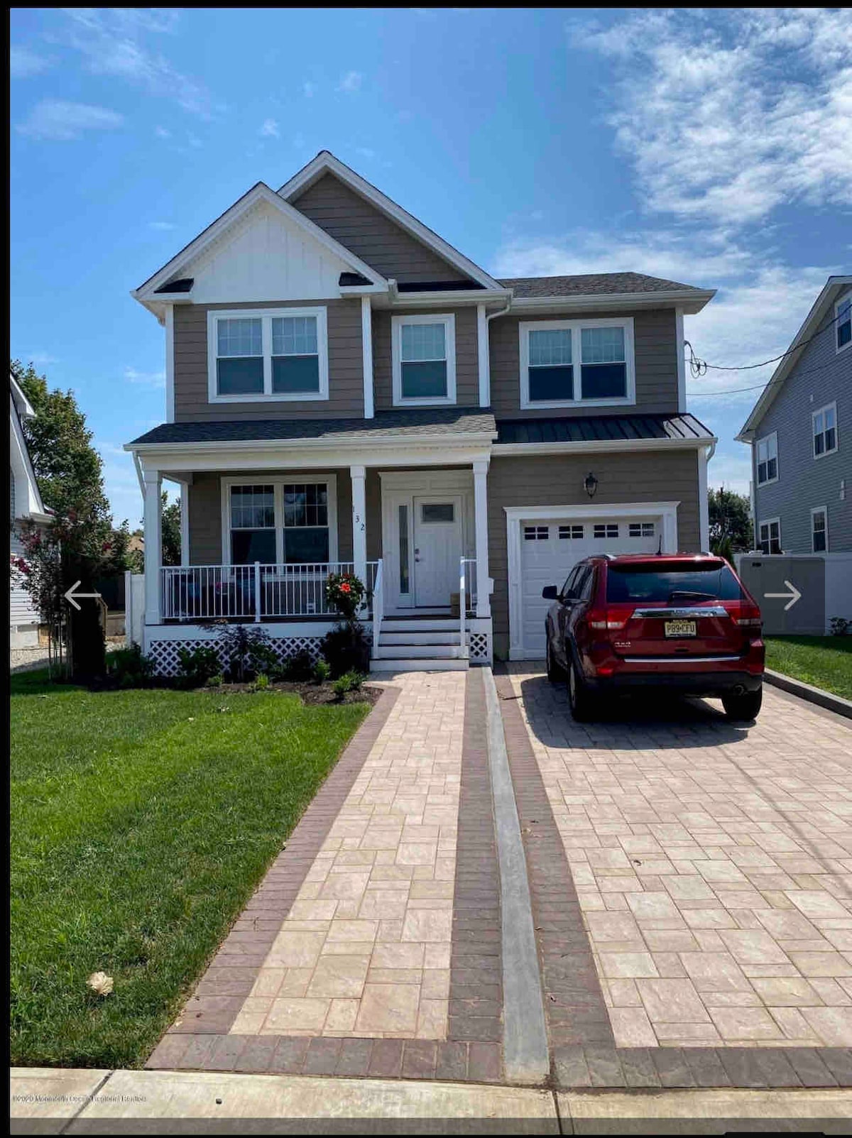 A two-story house features a light grey and white exterior with a front porch adorned with potted plants. A paved driveway accommodates a parked vehicle, while a small lawn adds greenery to the facade. Windows are symmetrically arranged on the upper level.