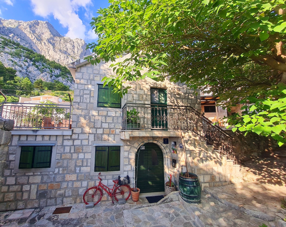 A charming stone house features a balcony above the entrance, surrounded by greenery. The exterior is complemented by a red bicycle and a decorative barrel near the door. The backdrop includes a rugged mountain landscape, showcasing the natural surroundings.