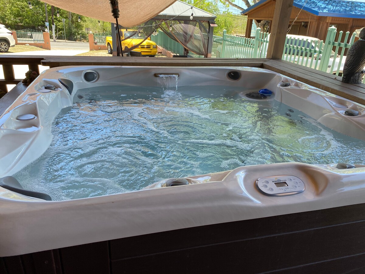 A spacious jacuzzi is filled with sparkling water, ready for relaxation. The surrounding wooden deck features a canopy providing shade, while hints of outdoor greenery and a colorful vehicle are visible in the background.