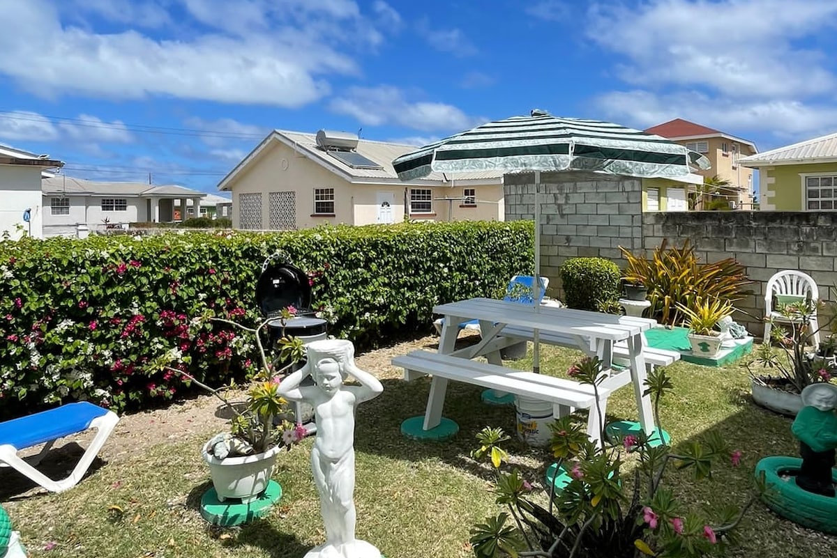 A vibrant outdoor area is highlighted by a white picnic table and chairs beneath a green striped umbrella. Lush greenery surrounds the space, including flowering plants and decorative statues. A clear blue sky complements the serene atmosphere of this well-maintained garden.