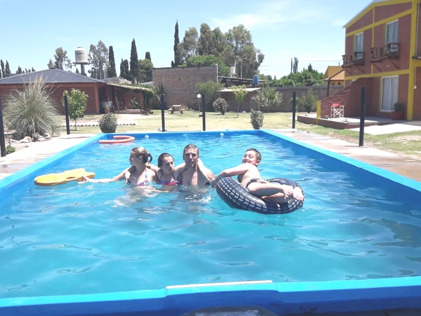 A swimming pool is shown, with children engaged in play. Colorful pool floats are visible, surrounded by a well-maintained outdoor area with greenery. Sunlight reflects off the water, enhancing the inviting atmosphere of this spacious outdoor setting.