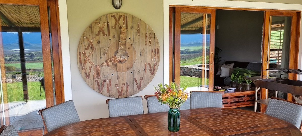 An inviting outdoor dining area features a large round table surrounded by multiple chairs. A wooden clock is mounted prominently on the wall, while sliding doors lead to an interior space where comfortable seating can be seen. Lush greenery and scenic views are visible beyond.