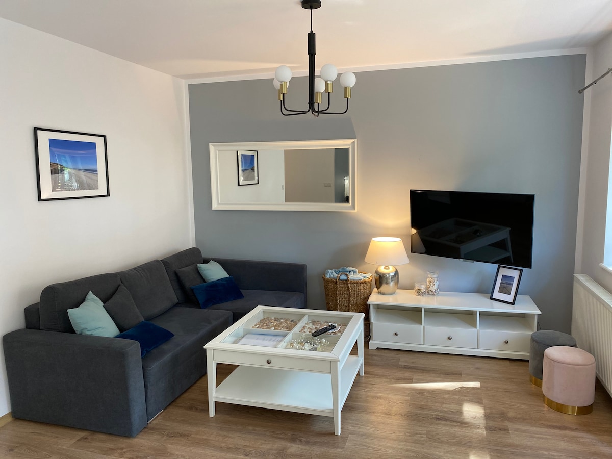 The living area features a comfortable gray sofa with decorative cushions. A white coffee table holds magazines, while a mirrored wall reflects the space. A television is mounted on the wall opposite a lamp, and artwork is displayed, enhancing the modern decor.