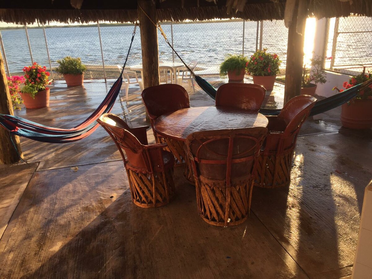A shaded outdoor seating area is furnished with a round table surrounded by four woven chairs. Colorful hammocks are suspended nearby, and potted plants with vibrant flowers are arranged around the space. The water's edge is visible in the background, reflecting soft light.