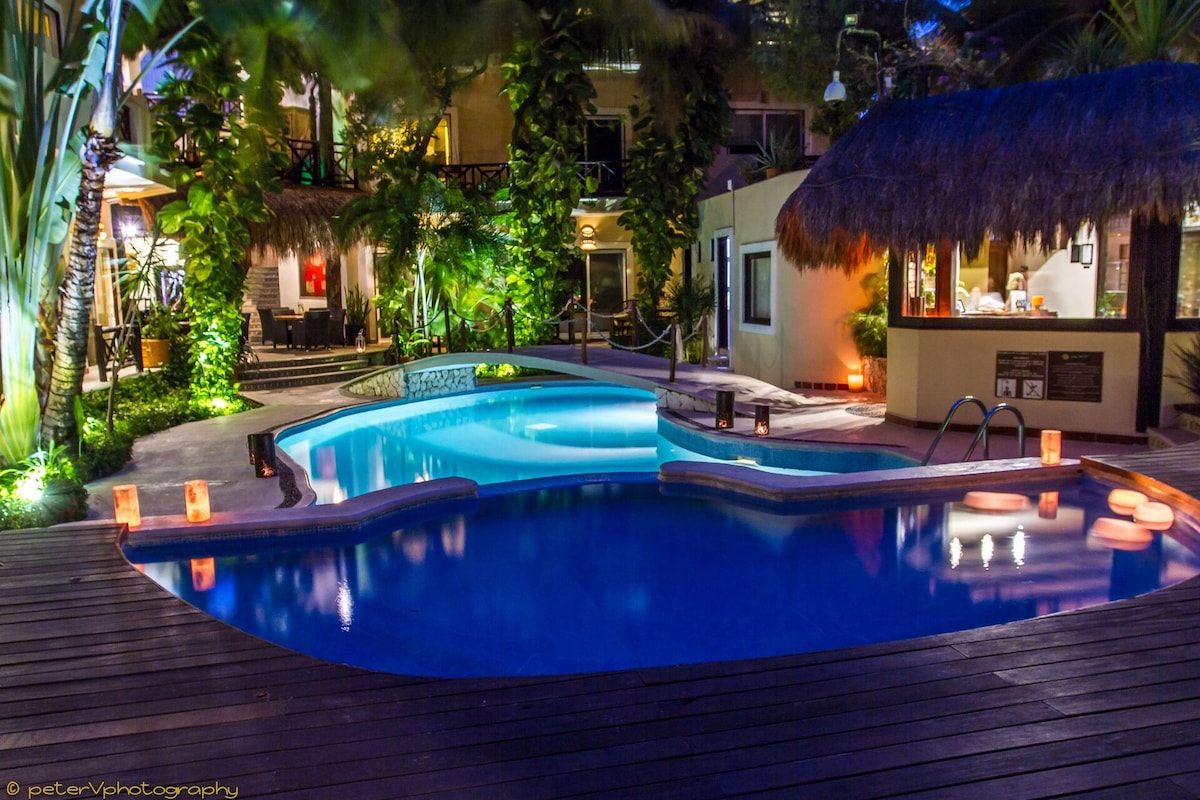 A tranquil swimming pool is surrounded by lush tropical plants and softly lit by lanterns, creating a serene environment. The pool features a gradual slope and multiple levels, inviting relaxation. A nearby bar area can be seen with a thatched roof, enhancing the outdoor experience.