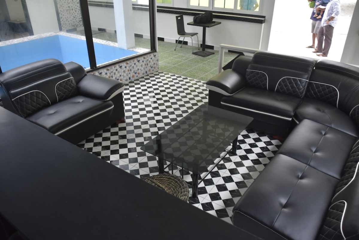 A modern seating area features a large sectional sofa with contrasting white stitching, paired with a glass coffee table. The floor showcases a checkered pattern of black and white tiles, and large windows provide a view of the nearby pool area.
