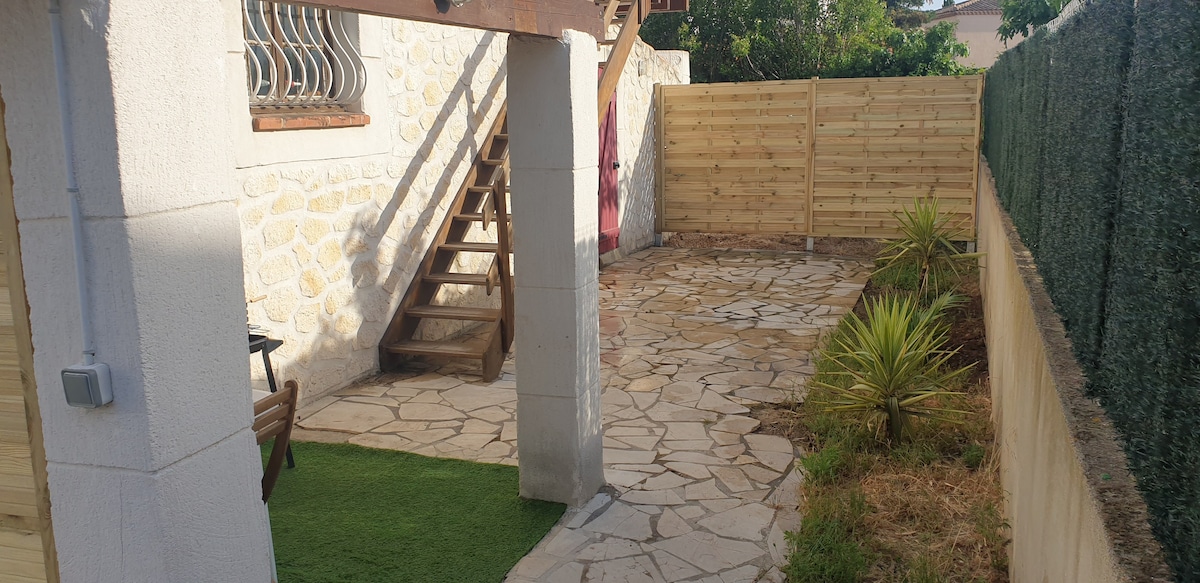 A private outdoor space features a patterned stone patio surrounded by greenery. A wooden staircase leads to an upper level, and a section of the yard has artificial grass. Fencing provides privacy, while a small palm plant adds a touch of nature.