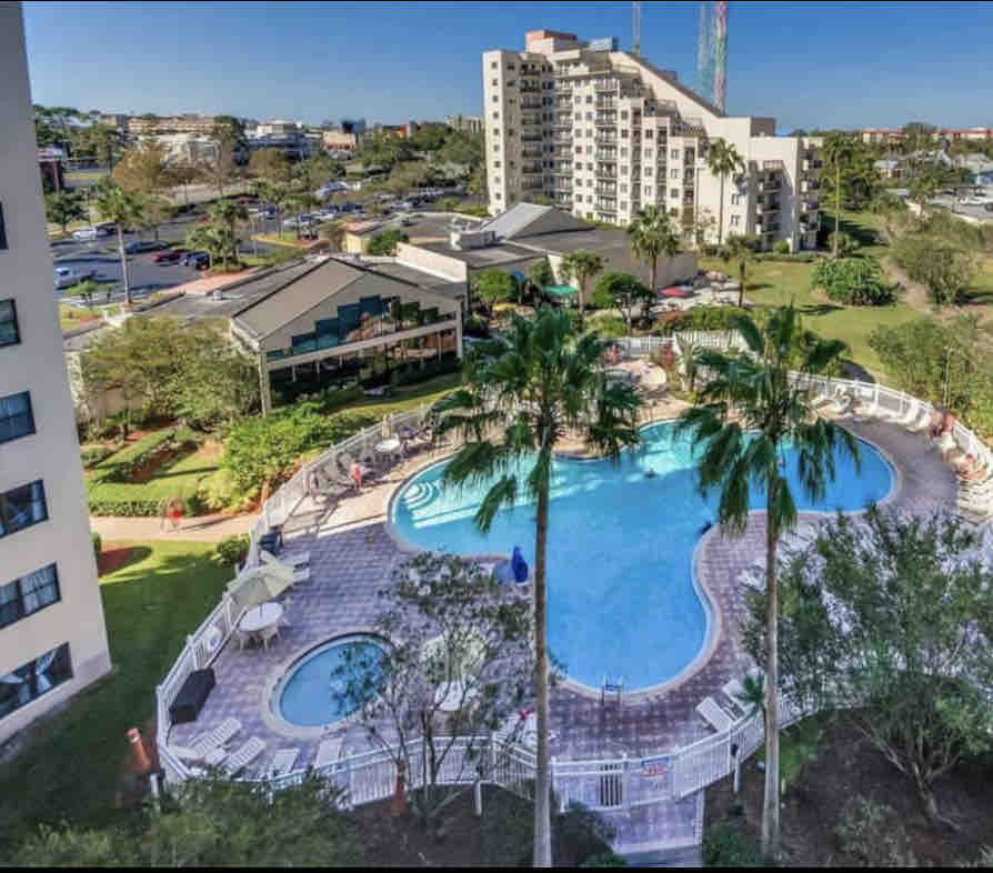 An aerial view presents two outdoor pools surrounded by lush landscaping and palm trees. Lounge chairs are positioned around the pool area, providing space for relaxation. A building is visible in the background, alongside vibrant greenery and open spaces.
