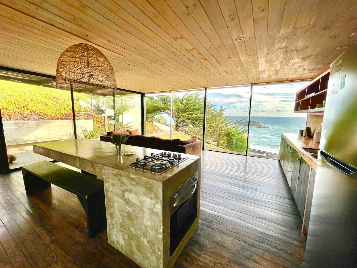 An open-concept kitchen and living area features a large concrete island with a gas stove. Floor-to-ceiling glass doors provide expansive views of the ocean and surrounding landscape. Natural light fills the space, enhancing the warm wooden interior elements and inviting seating arrangement.