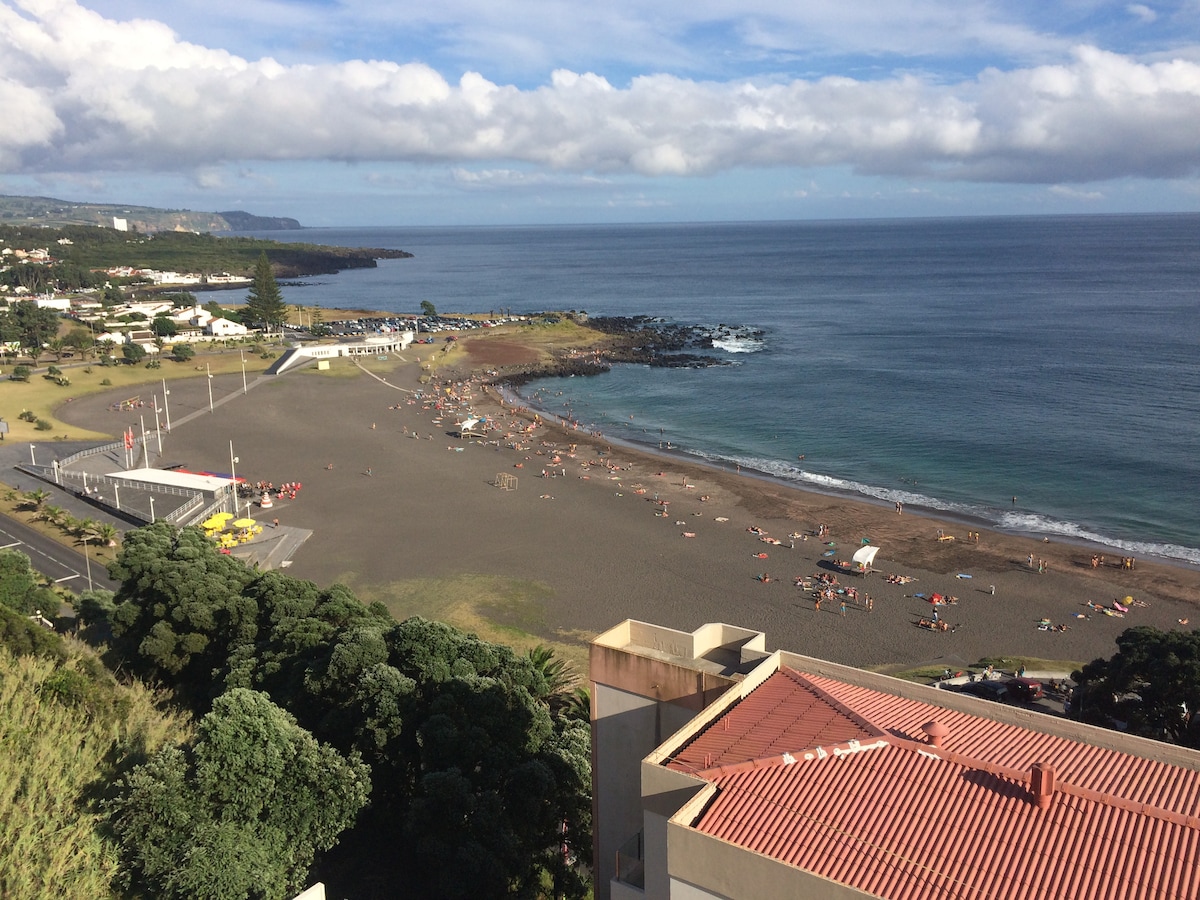 A scenic view of a beach is presented, featuring sandy shores and gentle waves. Several individuals are seen enjoying the sun and water. The beach is framed by lush greenery and coastal rocks, under a partly cloudy sky, highlighting the natural beauty of the area.