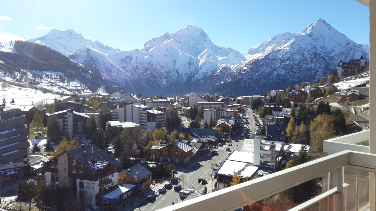 A panoramic view of snow-capped mountains is visible from the balcony, showcasing a vibrant village below. The layout of the area includes various buildings, trees, and streets, all set against a clear blue sky. Sunlight highlights the mountain peaks and the cozy atmosphere of the town.