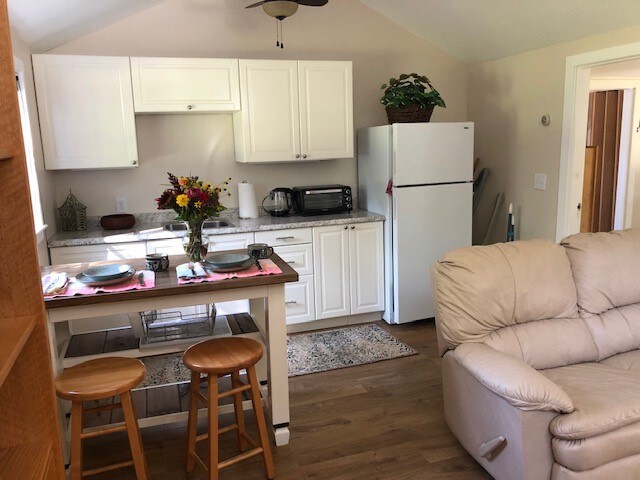 A well-equipped kitchen area is shown, featuring white cabinetry and a granite countertop. A full-size refrigerator stands adjacent to a microwave and toaster oven. Two wooden stools sit at a small table, which is set with place settings. A leather recliner is visible in the foreground.