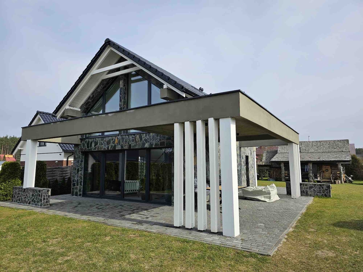 A modern two-story house features a striking design with large glass windows and a covered patio. The exterior showcases a combination of stone and wood elements, while the spacious surrounding grass area invites outdoor relaxation.
